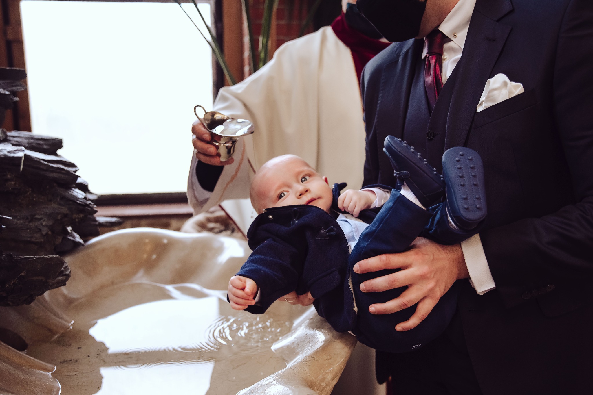 Niemowlę w granatowym ubranku trzymane nad marmurową chrzcielnicą podczas ceremonii, ksiądz w tle polewa wodą z metalowej miseczki.