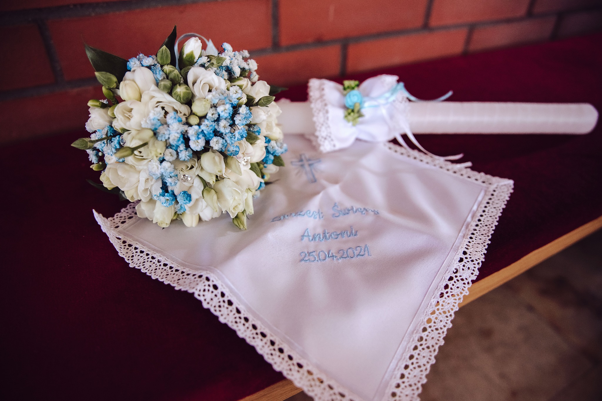 Kompozycja na Chrzest Święty: bukiet białych kwiatów z niebieskimi akcentami, świeca z dekoracją i haftowana chusteczka z imieniem Antoni i datą 25.04.2021.