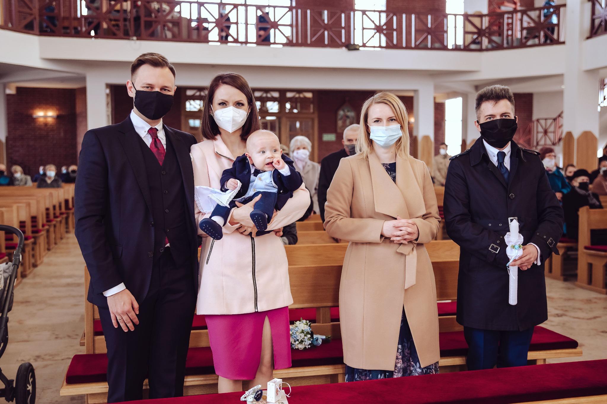 Rodzina w kościele, ubrana elegancko i w maseczkach, podczas uroczystości, prawdopodobnie chrztu, mężczyzna trzyma świecę.