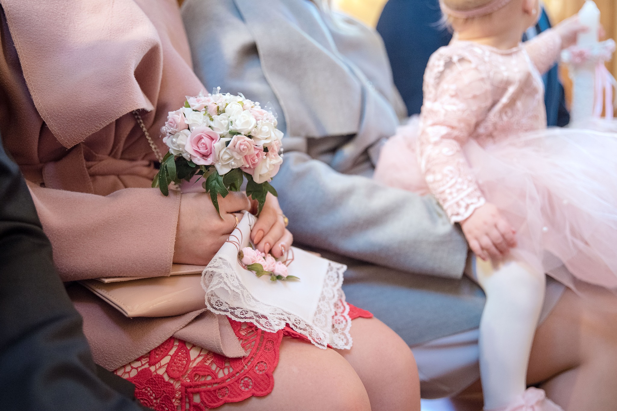 Uczestnicy uroczystości: kobieta trzyma bukiecik białych i różowych róż oraz ozdobną chusteczkę, obok siedzi mała dziewczynka w tiulowej sukience z dekoracyjną świecą.