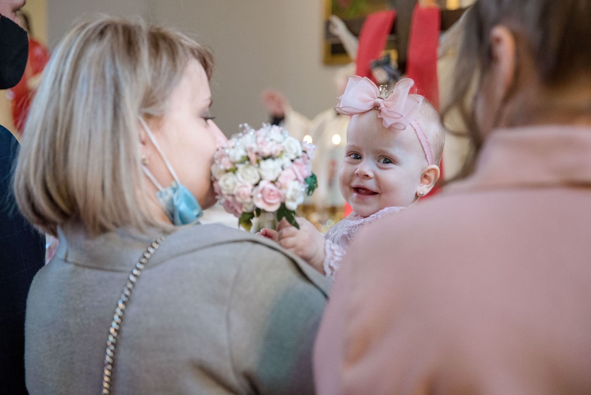 Uśmiechnięta dziewczynka z różową kokardą na głowie, trzyma bukiet kwiatów podczas uroczystości, widoczna mama w maseczce i fragment kościoła w tle.