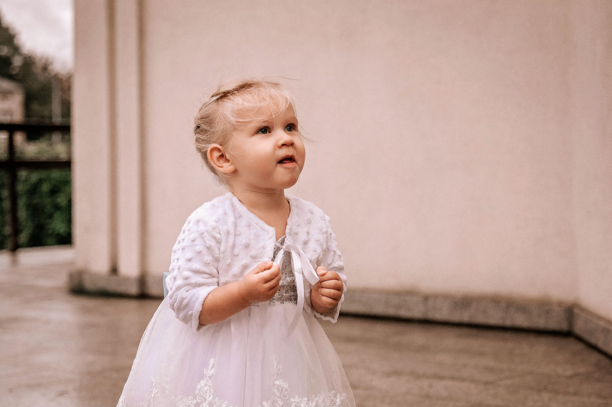 Mała dziewczynka w białej sukience i bolerku spogląda w górę z lekkim zaciekawieniem, delikatne włosy opadają na twarz, tło stanowią jasne filary i ściana.