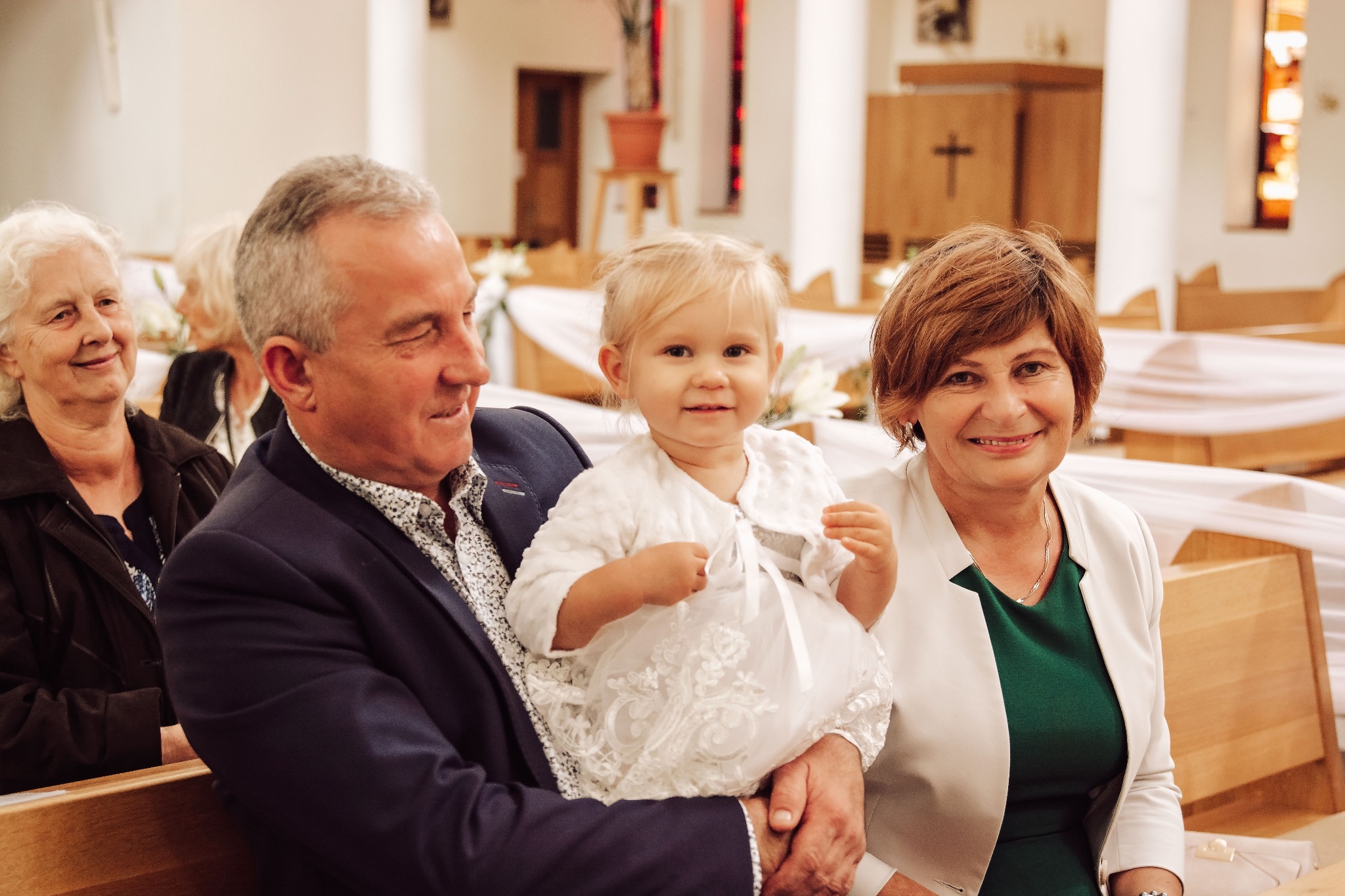 Uśmiechnięta dziewczynka w białej sukience trzymana na kolanach przez mężczyznę w kościele, obok siedzą dwie uśmiechnięte kobiety, dekoracje kwiatowe w tle.