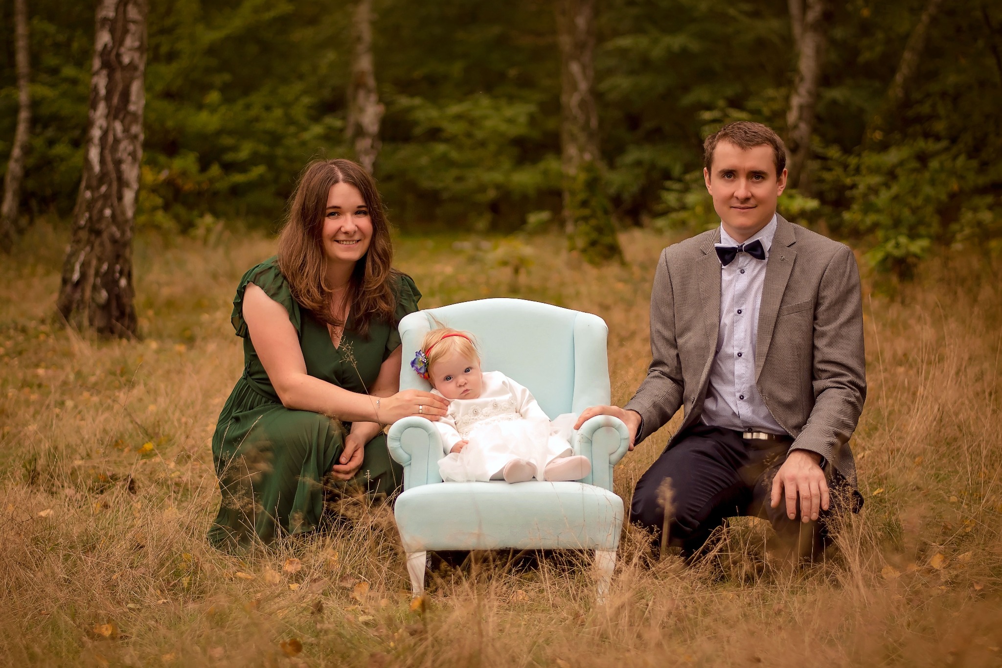 Rodzinna sesja zdjęciowa w plenerze: rodzice klęczą obok małego dziecka w białej sukience siedzącego na jasnoniebieskim fotelu w otoczeniu jesiennej trawy i drzew.