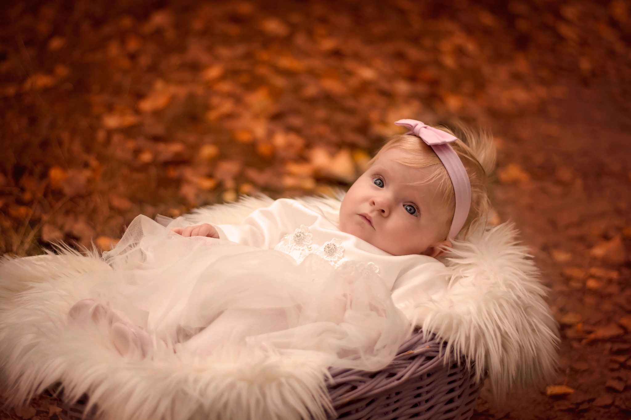 Niemowlę w białej sukience i różowej opasce leży w koszu wyłożonym białym futrem, na tle jesiennych liści.