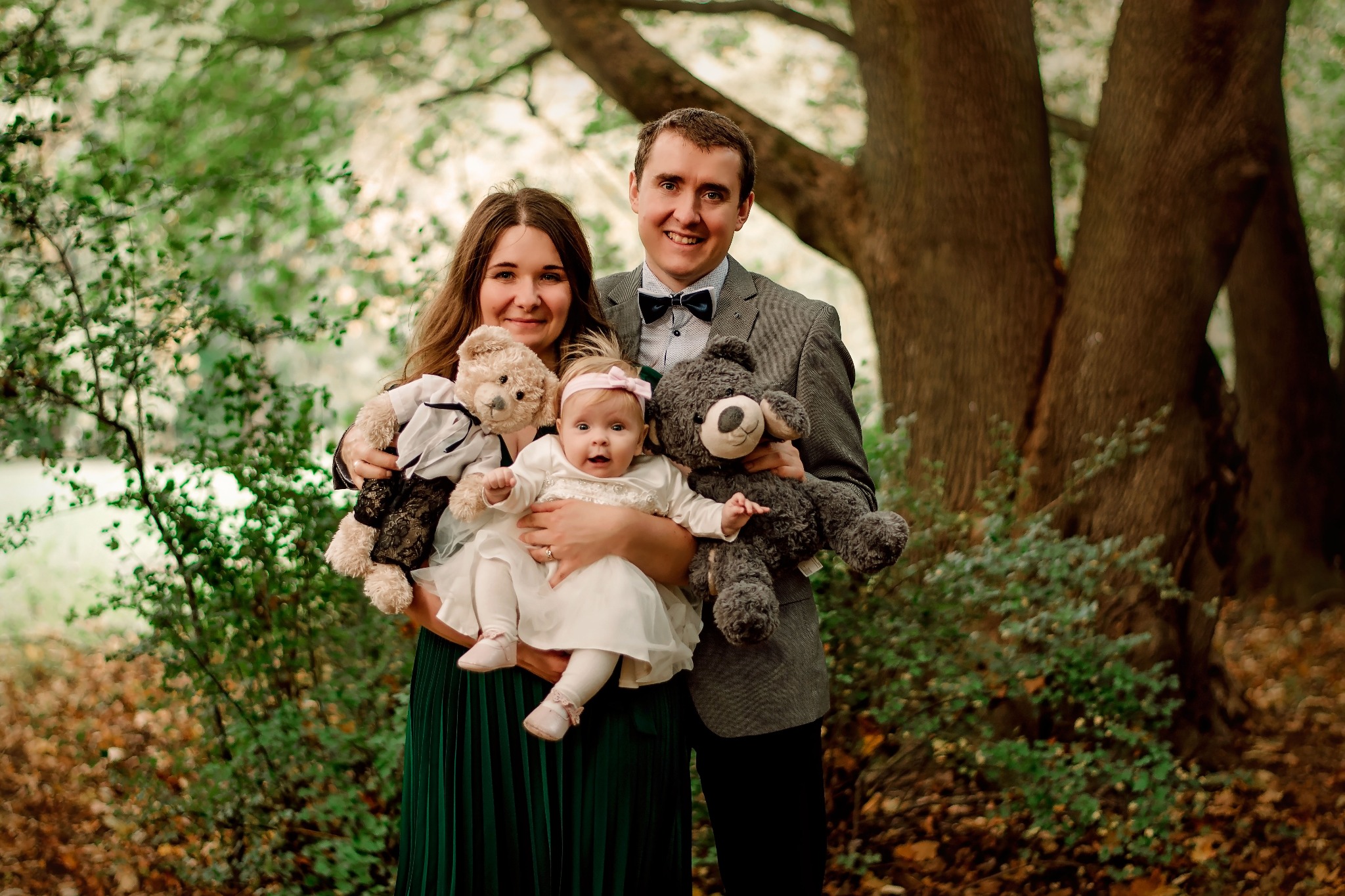 Rodzinne zdjęcie w plenerze: rodzice trzymają dziecko i dwa pluszowe misie na tle drzew i zieleni.