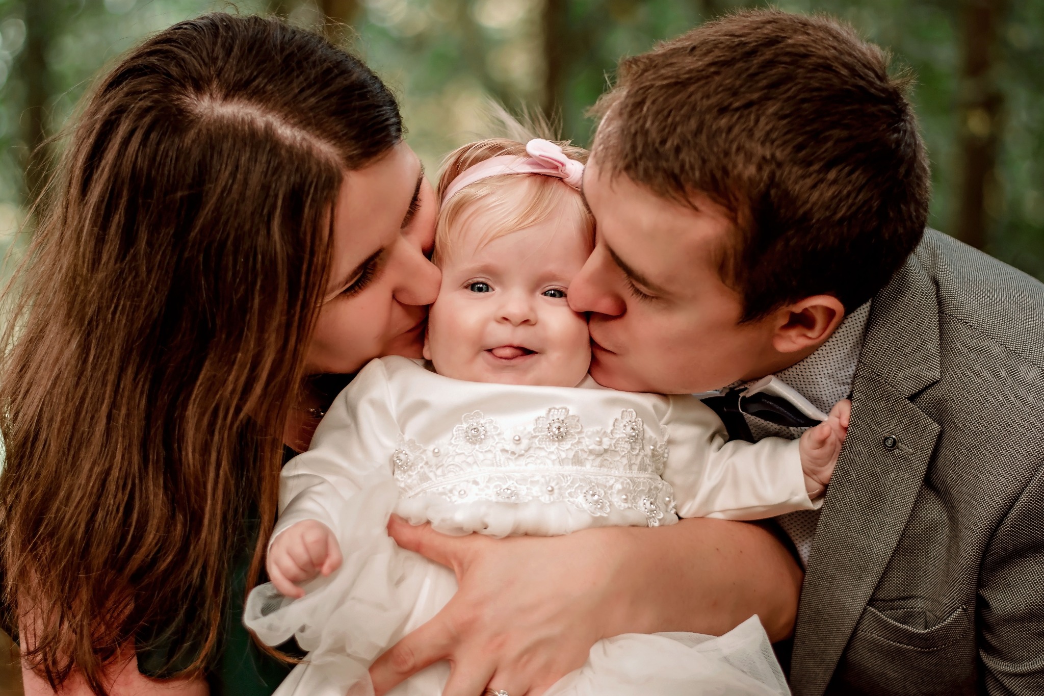 Rodzice całują w policzki uśmiechnięte niemowlę w białej sukience z kokardką na tle zieleni.