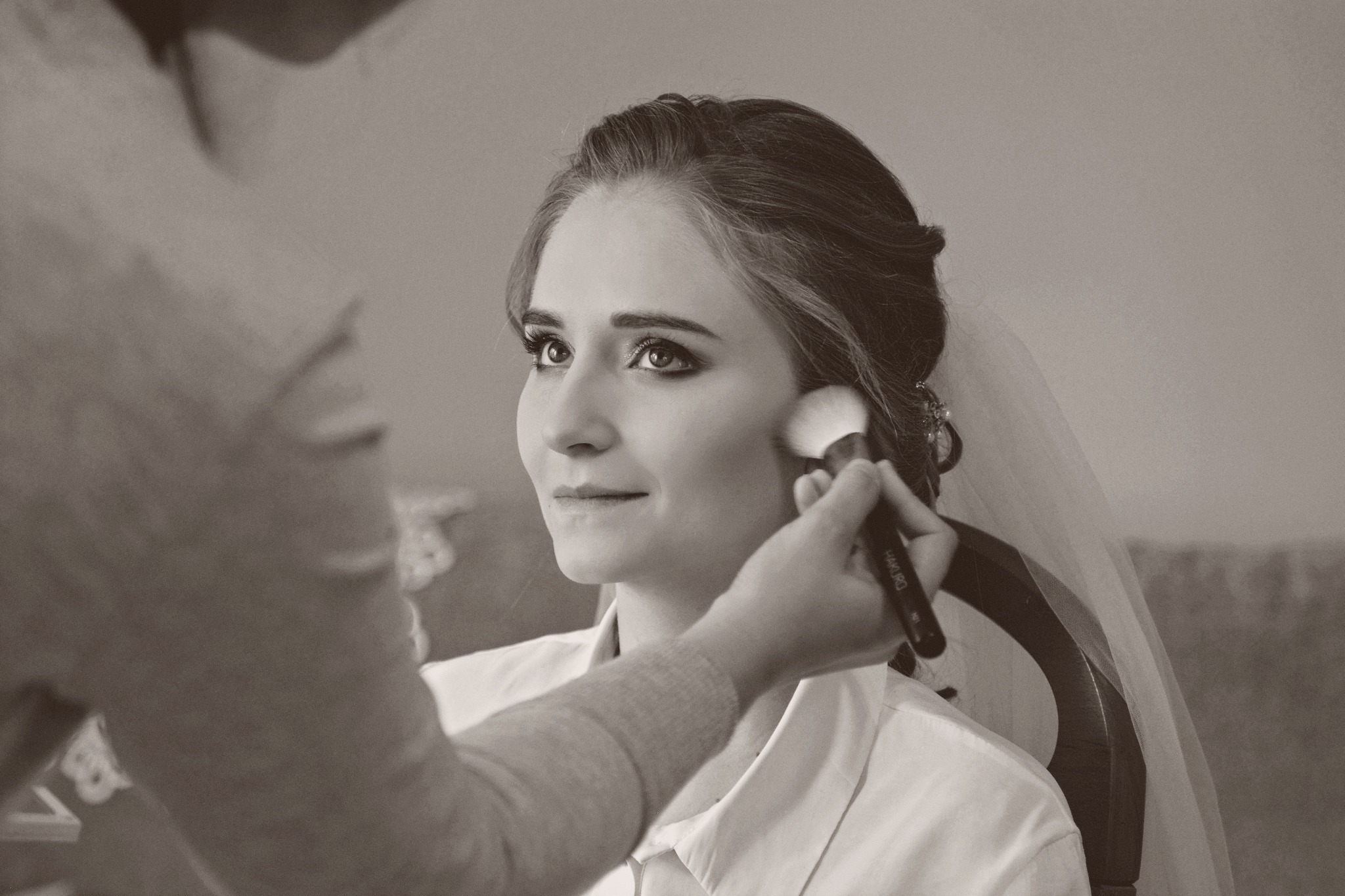 Panna młoda w trakcie makijażu ślubnego, wizażystka aplikuje puder pędzlem Hakuro, widoczny welon i upięcie włosów.