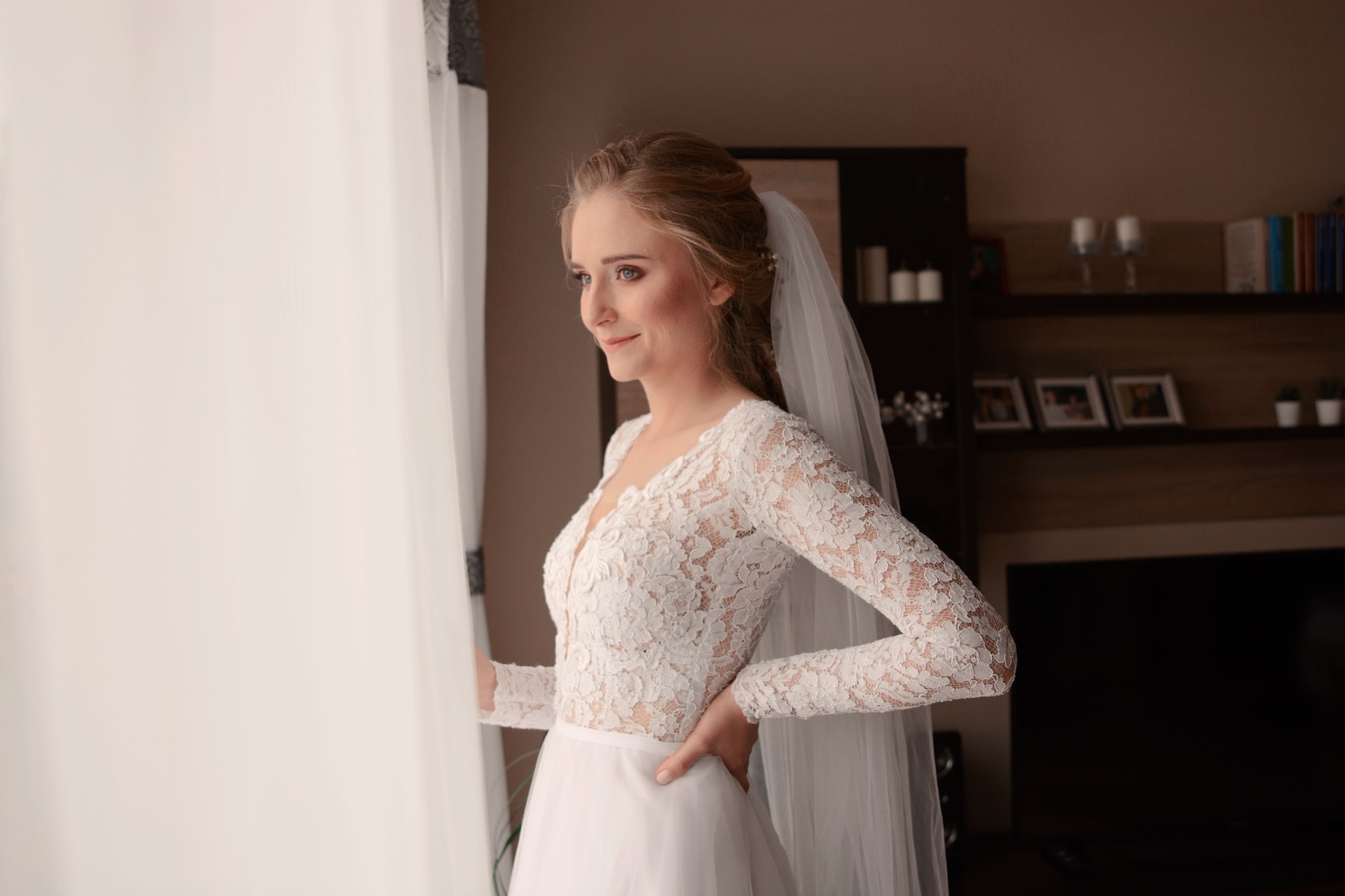 Panna młoda w koronkowej sukni z długim rękawem i welonem, spogląda przez okno, trzymając rękę na biodrze, w tle regał z książkami i zdjęciami.