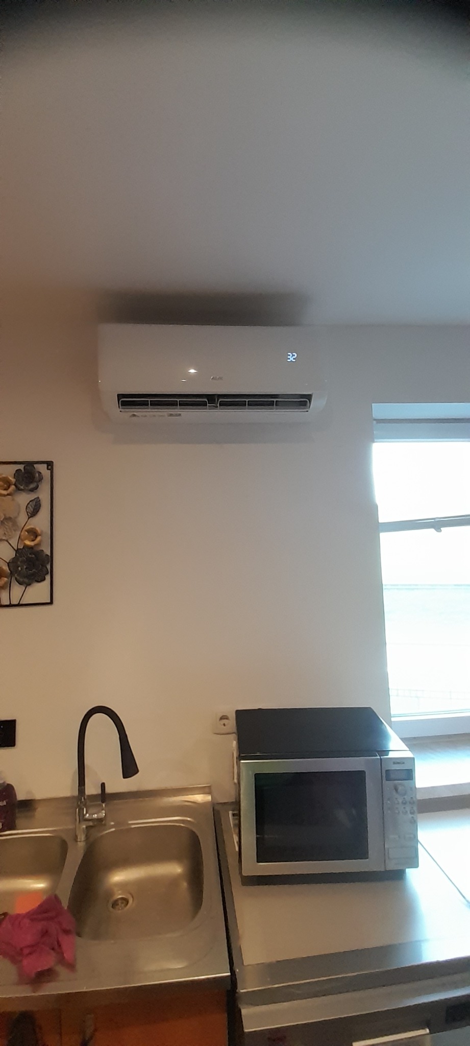 Biały klimatyzator ścienny z wyświetlaczem temperatury 32 stopnie Celsjusza, zamontowany w kuchni nad zlewem i mikrofalówką, obok okna.
