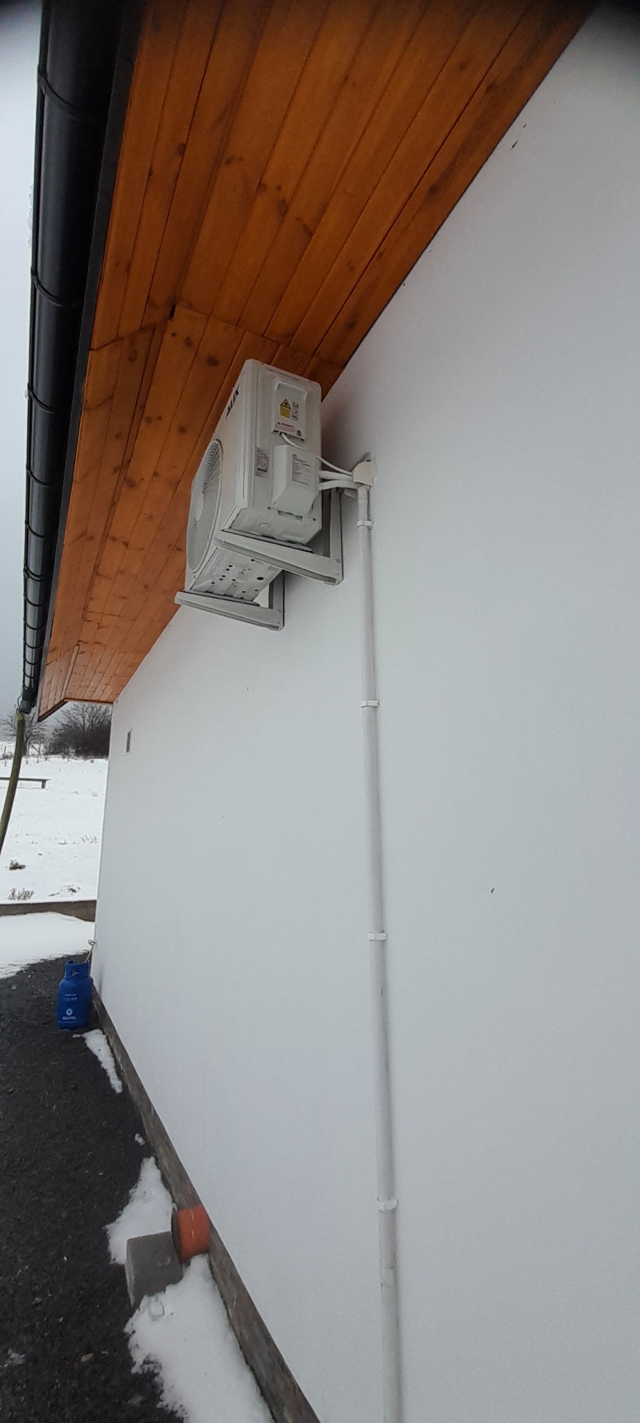 Zewnętrzna jednostka klimatyzacji marki AUX zamontowana na białej ścianie budynku z drewnianym podbitką, widoczna instalacja odprowadzająca skropliny. Na ziemi leży śnieg.