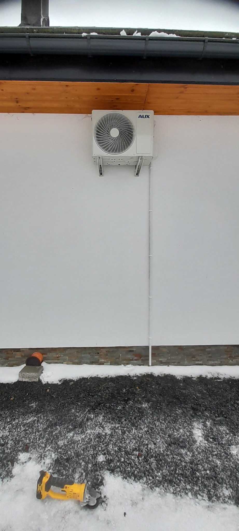 Zamontowana jednostka zewnętrzna klimatyzacji AUX na białej ścianie budynku z widocznymi przewodami. Na ziemi leży szlifierka kątowa DeWalt.