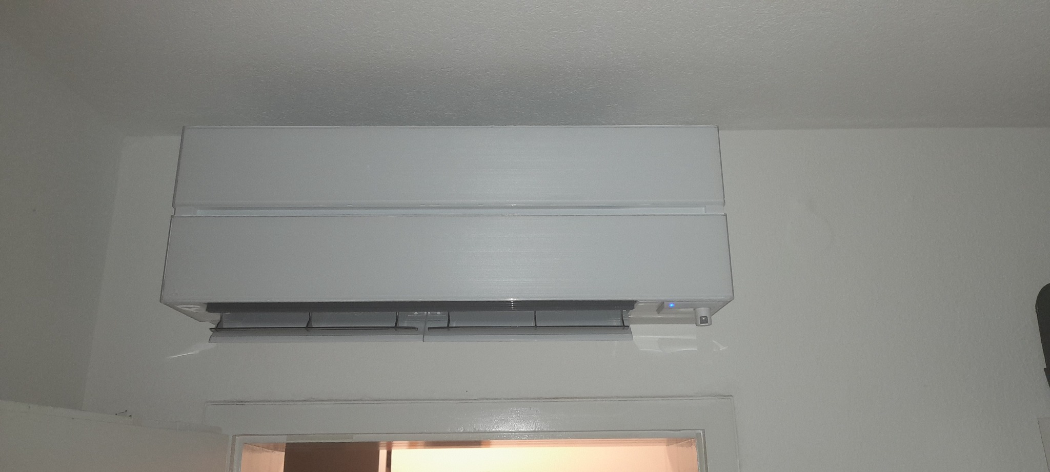 Biały klimatyzator ścienny zamontowany nad futryną drzwi w pomieszczeniu mieszkalnym, widoczna niebieska dioda sygnalizacyjna.