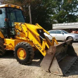 Żółta koparko-ładowarka marki JCB z łyżką z przodu, zaparkowana na ziemi, z widocznymi oponami z bieżnikiem i kabiną operatora, w tle samochód osobowy i ciężarowy.