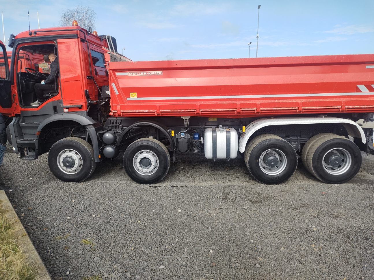 Czerwona wywrotka marki Meiller-Kipper z otwartą kabiną, widoczna na żwirowym podłożu, z osobą siedzącą wewnątrz.