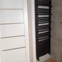 Czarny, drabinkowy grzejnik łazienkowy zamontowany na szarej ścianie obok białych drzwi z poziomymi wstawkami w nowoczesnej łazience z drewnopodobną podłogą.