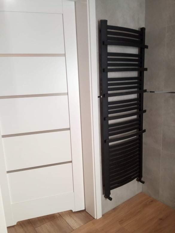 Czarny, drabinkowy grzejnik łazienkowy zamontowany na szarej ścianie obok białych drzwi z poziomymi wstawkami w nowoczesnej łazience z drewnopodobną podłogą.