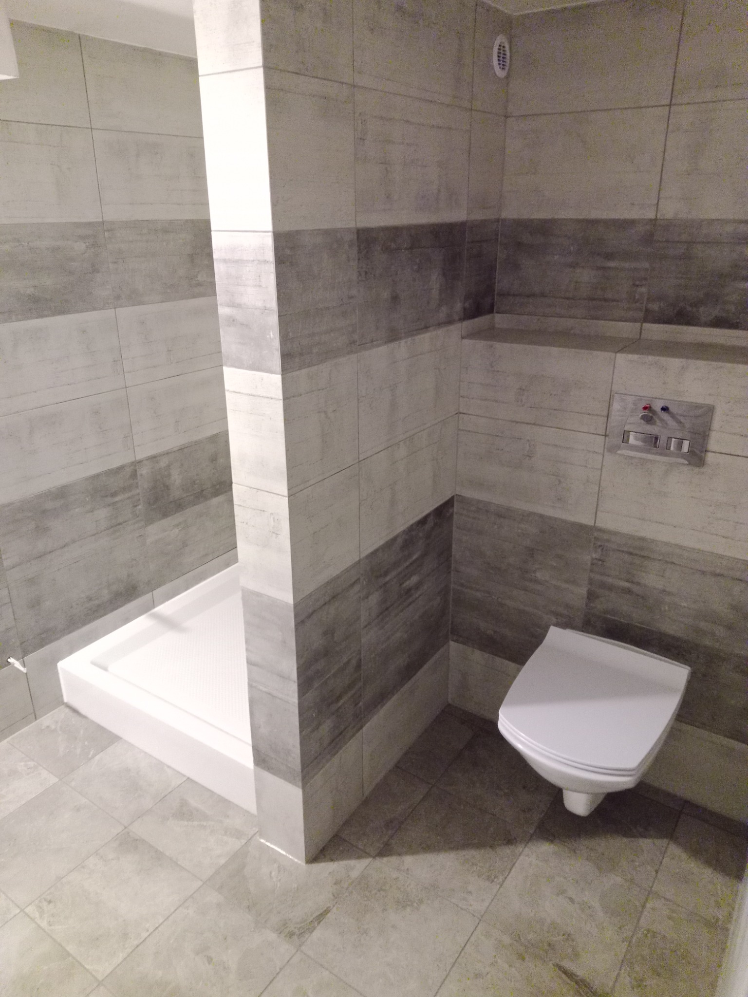 Wnętrze łazienki z kabiną prysznicową i podwieszaną toaletą, wykończone płytkami w odcieniach szarości i bieli.