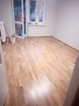 Puste pomieszczenie po remoncie z nową, jasną, drewnianą podłogą parkietową ułożoną w jodełkę, widoczne otwarte drzwi balkonowe i fragment grzejnika.