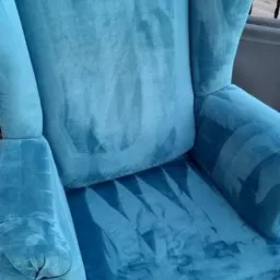 Turkusowy fotel typu uszak z widocznymi śladami po czyszczeniu tapicerki, na tle balkonu i dachu, z dyszą odkurzacza piorącego na pierwszym planie.