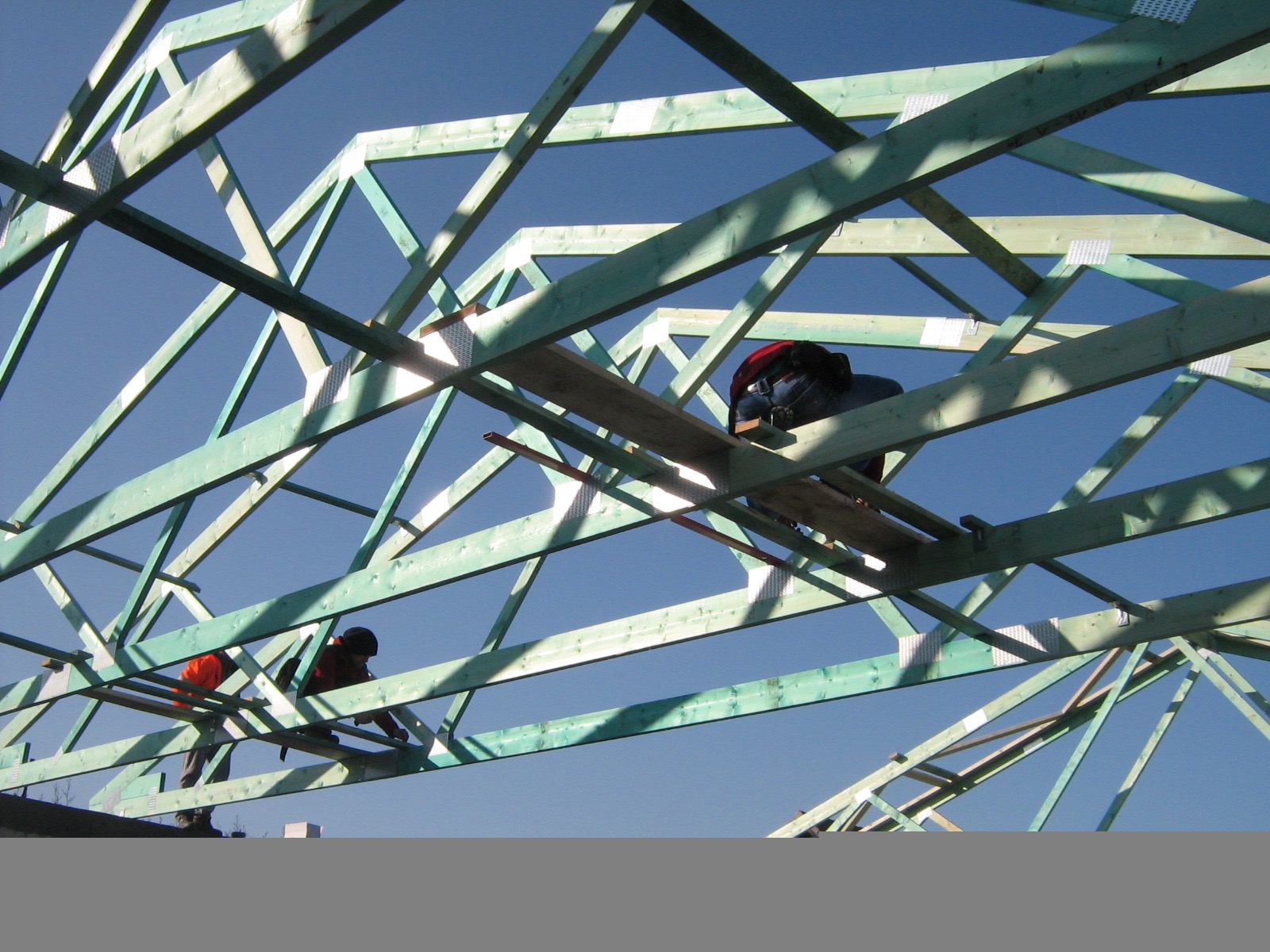 Konstrukcja drewniana dachu w trakcie montażu, dwóch robotników w uprzężach pracuje na wysokości, błękitne niebo w tle.