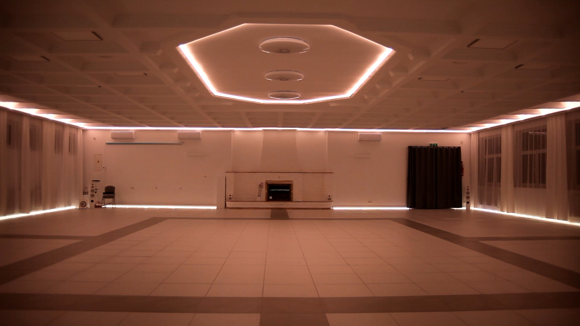 Duża, pusta sala z nowoczesnym oświetleniem LED na suficie w kształcie ośmiokąta, podświetlane listwy LED przy podłodze i suficie, białe ściany, okna z białymi zasłonami, parkiet podzielony...