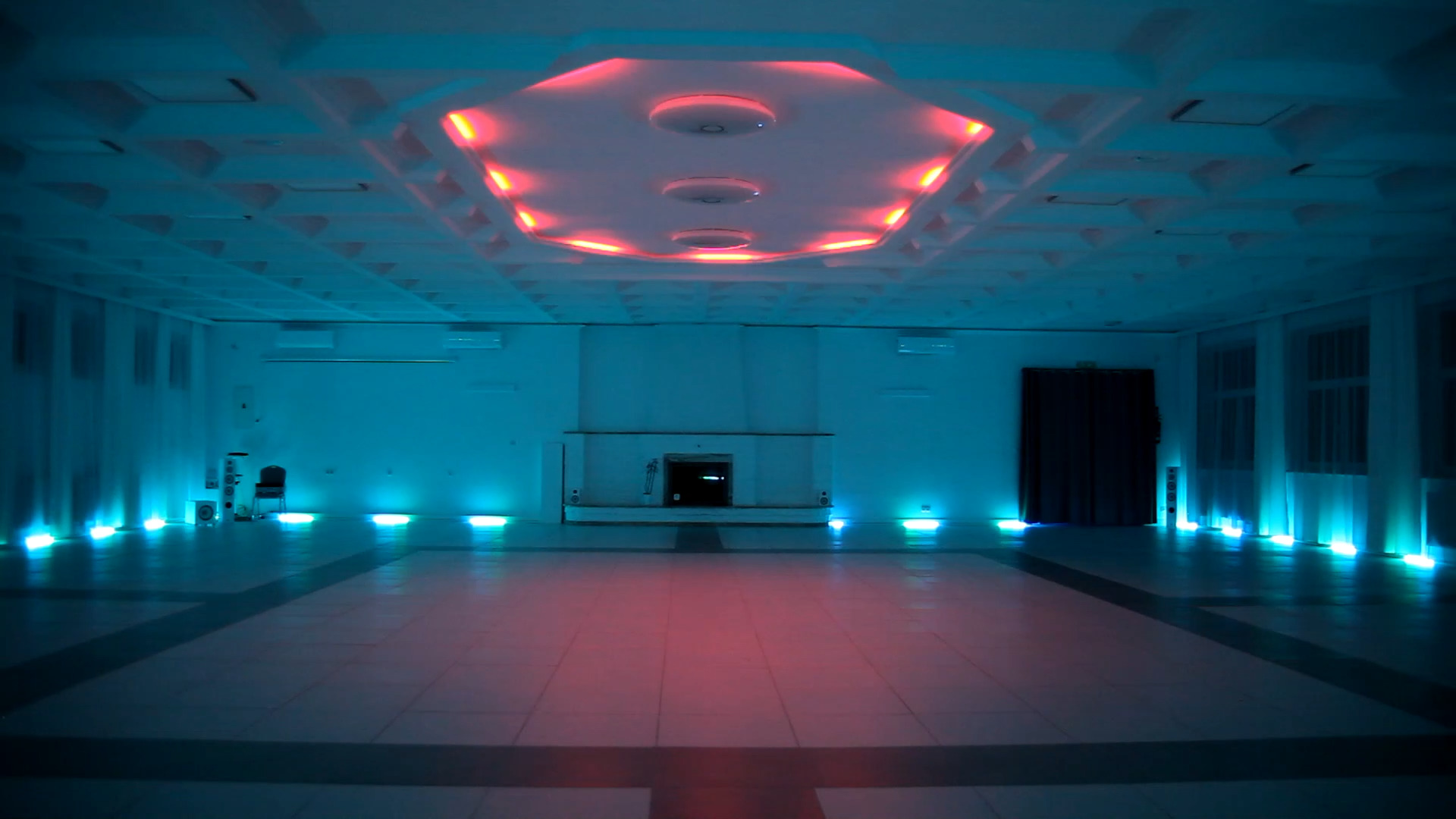 Pusta sala z podświetlanym sufitem kasetonowym i podłogą, oświetlenie w odcieniach niebieskiego i czerwonego, widoczny kominek i zasłonięte okna.