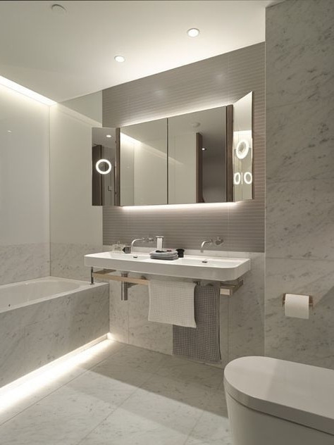 Nowoczesna łazienka z białymi marmurowymi płytkami, podświetlaną wanną i lustrem z wbudowanym pierścieniem świetlnym nad podwójną umywalką.