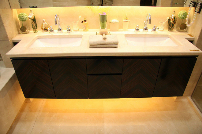 Nowoczesna łazienka z podwójną umywalką na blacie z jasnego kamienia, szafka z ciemnego drewna z wzorem w jodełkę, ozdobne flakoniki i rośliny na blacie, podświetlenie LED pod szafką.