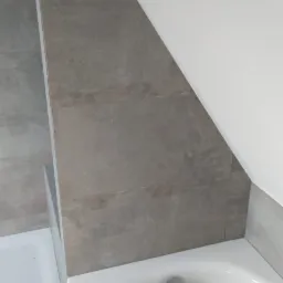 Szara płytka imitująca kamień na ścianie w łazience ze skosem, biała wanna akrylowa z odpływem, fragment brodzika z odpływem w rogu.