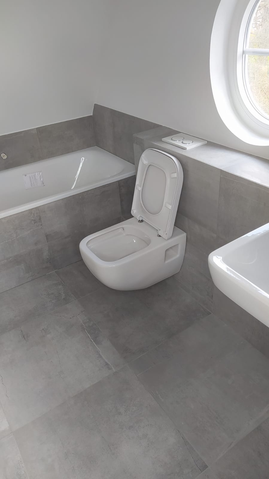 Nowoczesna łazienka z szarymi płytkami, wiszącą toaletą z otwartą klapą, białą wanną i umywalką, okrągłe okno w tle.