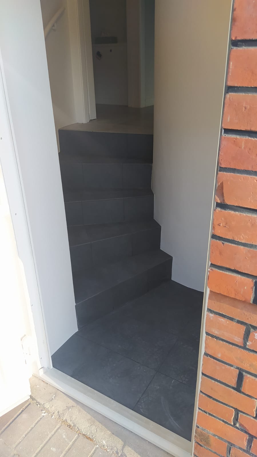 Wyłożone ciemnoszarą płytką schody wejściowe do domu, widoczne z zewnątrz, z fragmentem muru z czerwonej cegły po prawej stronie.