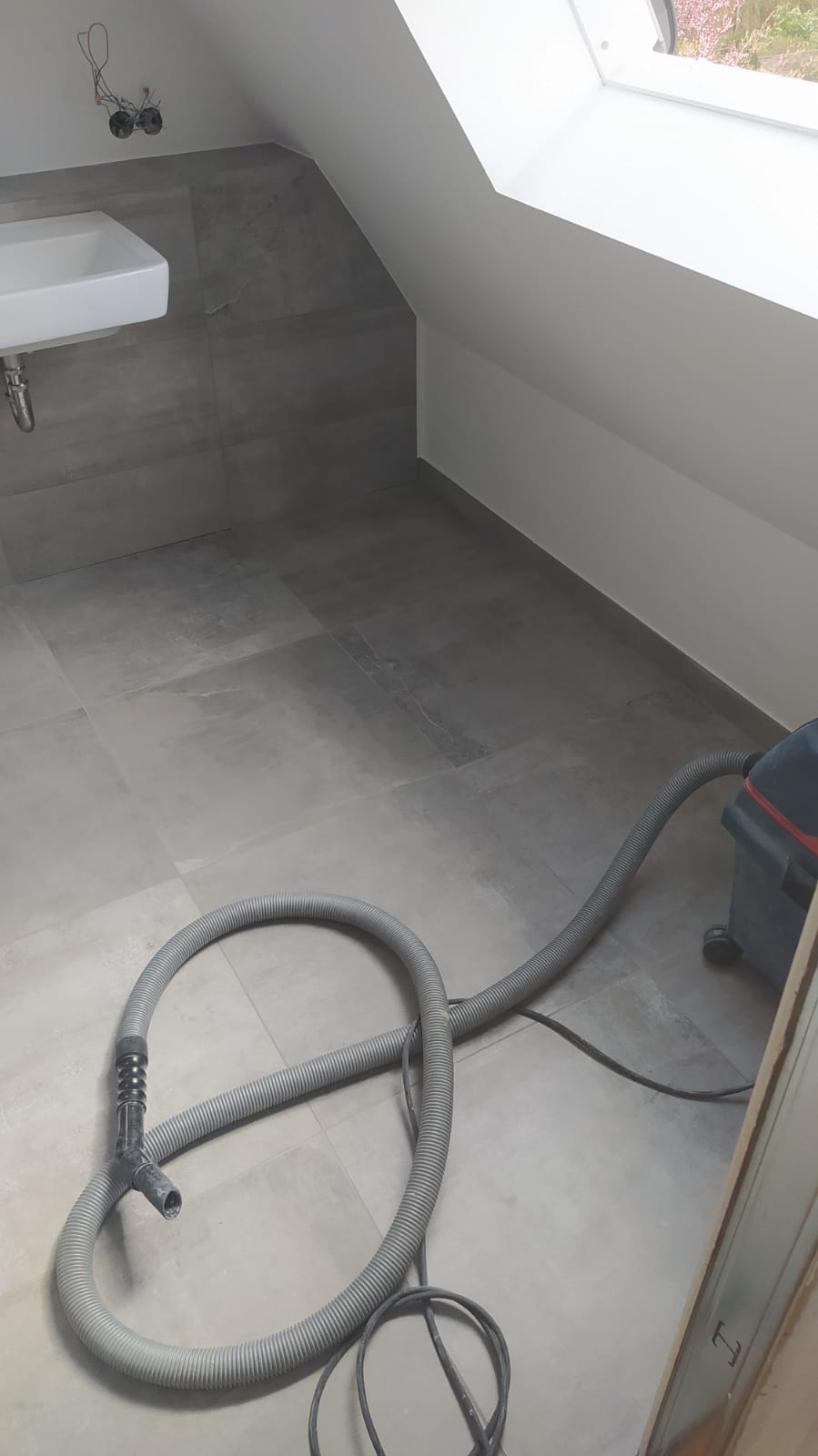 Szara łazienka na poddaszu z umywalką, szarymi płytkami i odkurzaczem przemysłowym podczas sprzątania po remoncie.