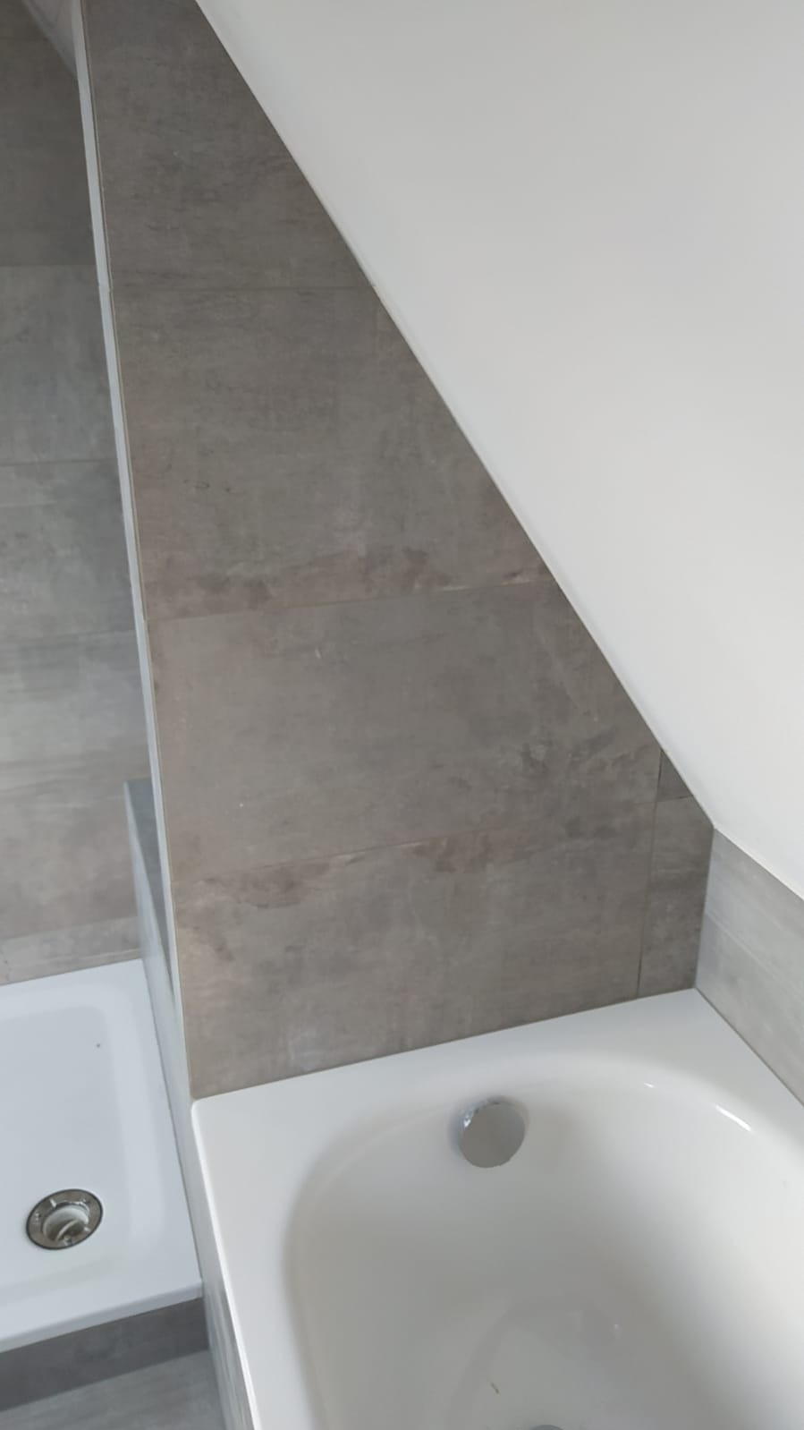 Szara płytka imitująca kamień na ścianie w łazience ze skosem, biała wanna akrylowa z odpływem, fragment brodzika z odpływem w rogu.