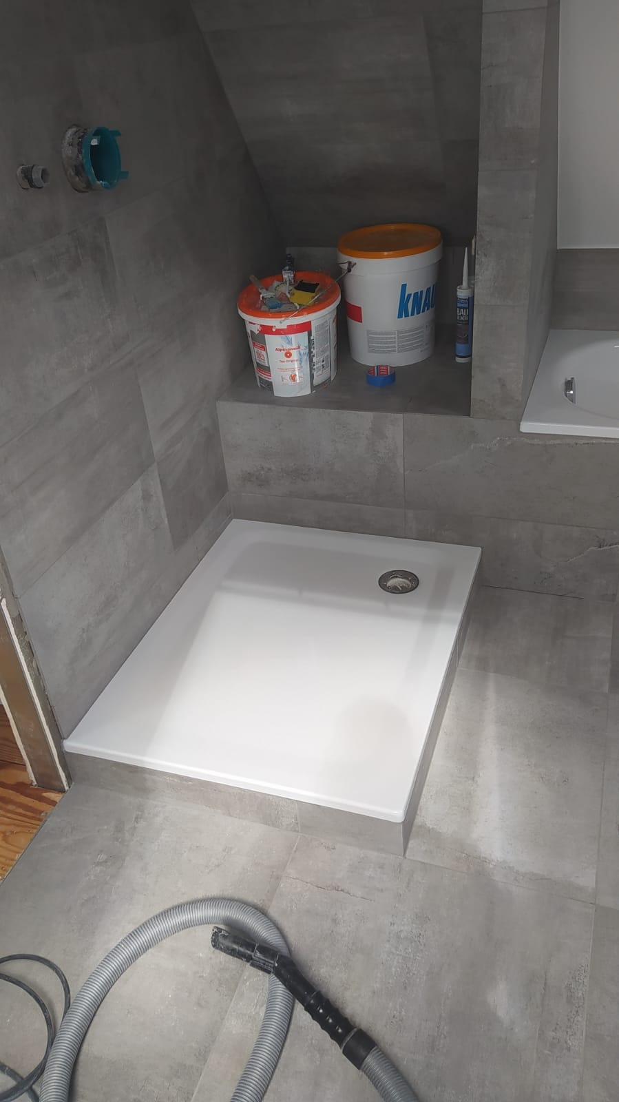 Montaż brodzika w łazience z szarymi płytkami, widoczne wiaderka z zaprawą i rura od odkurzacza na podłodze.
