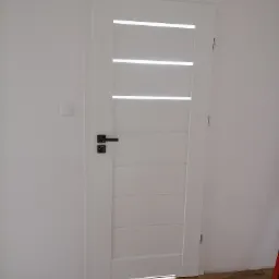 Białe drzwi wewnętrzne z poziomymi przeszkleniami i czarną klamką, zamontowane w białej ścianie nad panelami podłogowymi.