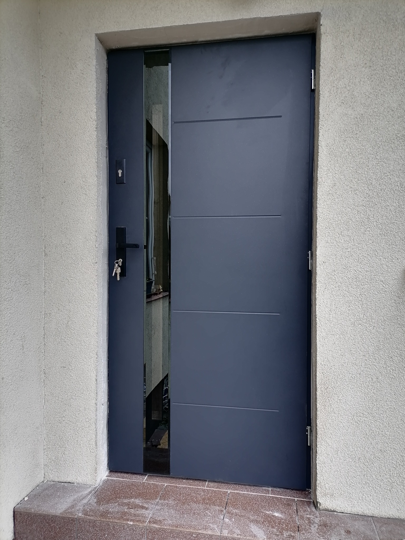 Nowoczesne, antracytowe drzwi wejściowe z przeszkleniem bocznym, widoczne klucze w zamku, ściana zewnętrzna z szorstkim tynkiem.