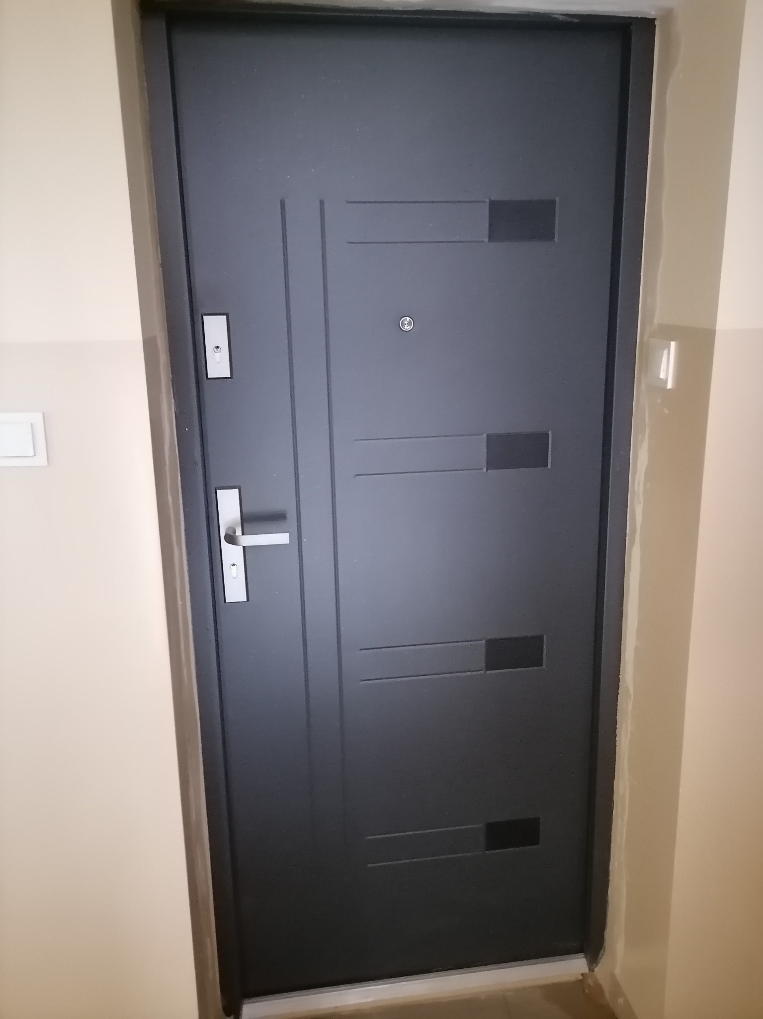 Ciemnoszare, nowoczesne drzwi wejściowe z poziomymi wgłębieniami i srebrną klamką, zamontowane w jasnym przedpokoju.