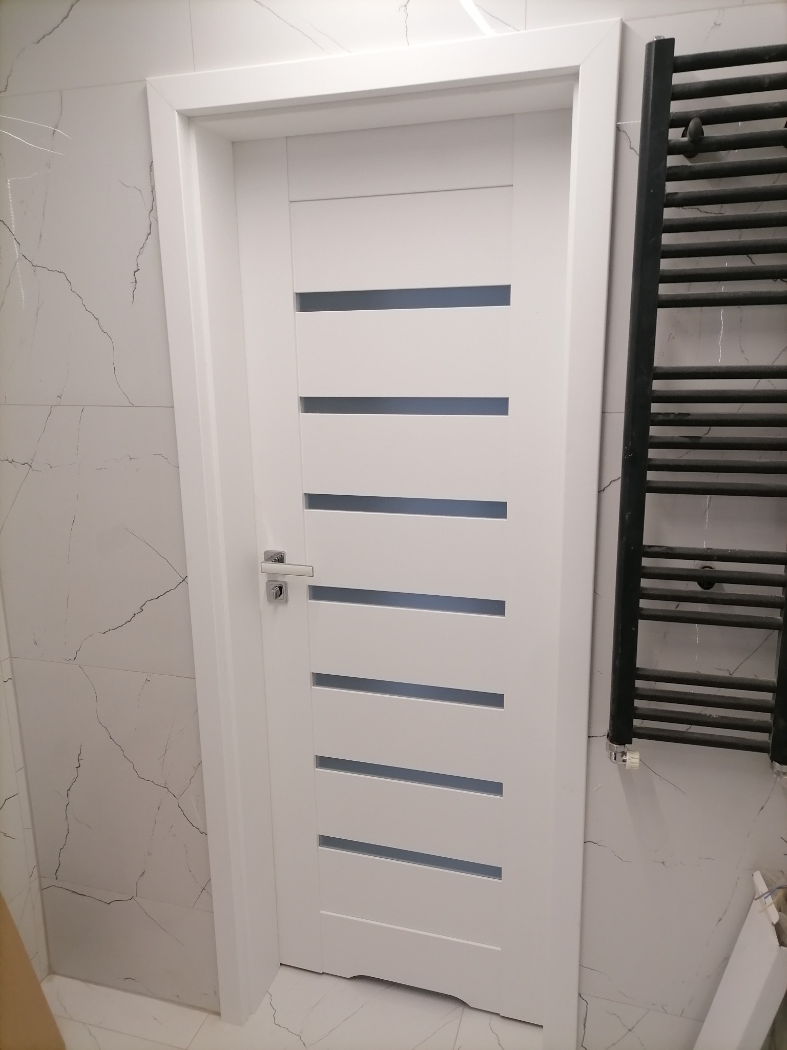 Białe drzwi wewnętrzne z poziomymi przeszkleniami i chromowaną klamką, zamontowane w łazience z marmurową okładziną ścienną, obok grzejnik drabinkowy.