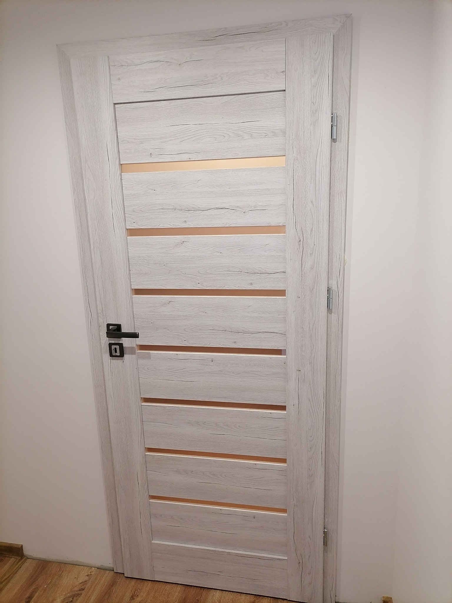 Zamknięte, wewnętrzne drzwi w odcieniach szarości z poziomymi, transparentnymi paskami, czarna klamka i widoczny fragment parkietu.