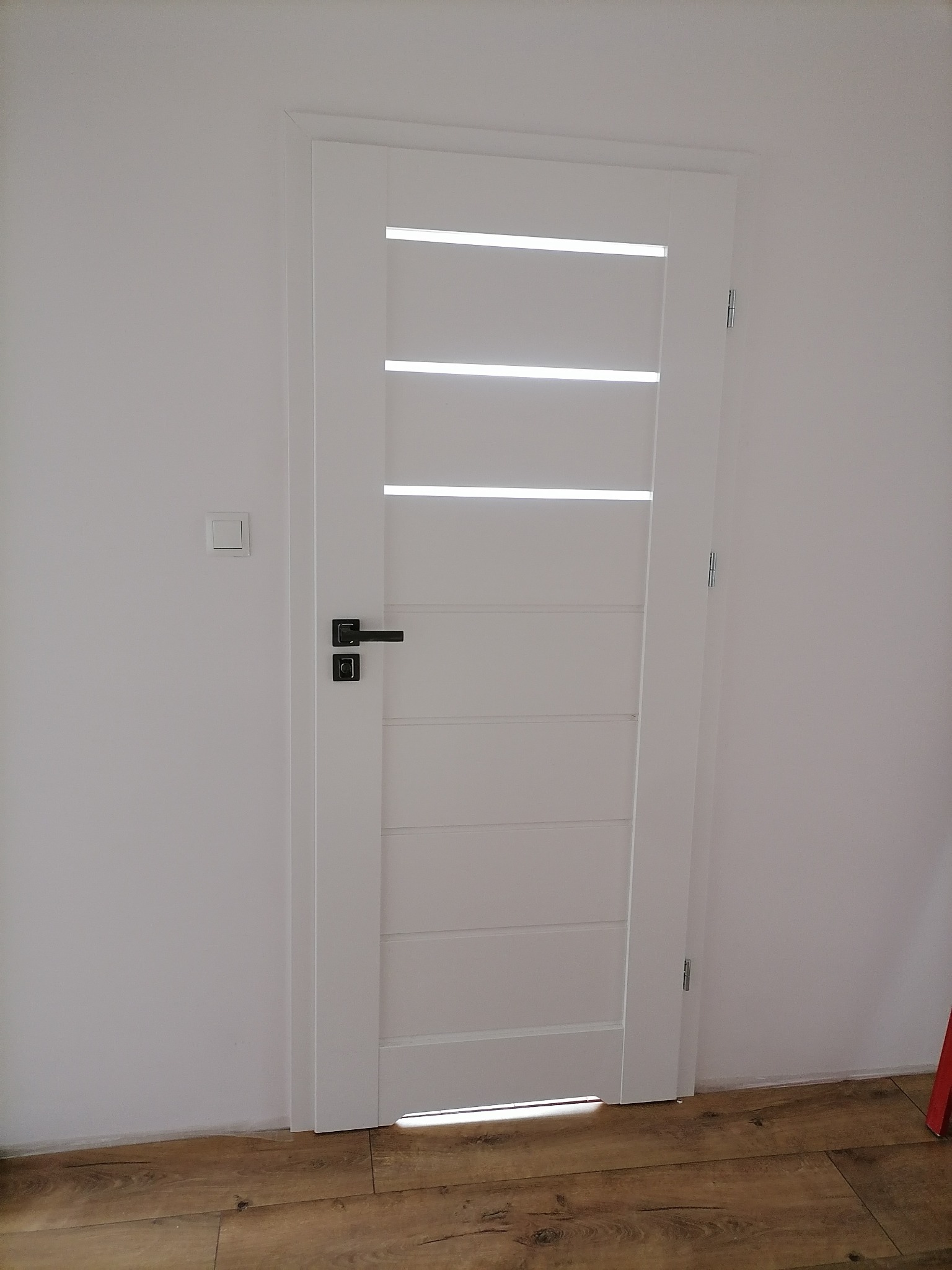 Białe drzwi wewnętrzne z poziomymi przeszkleniami i czarną klamką, zamontowane w białej ścianie nad panelami podłogowymi.