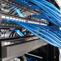 Szafa rack z zainstalowanymi przełącznikami PoE marki BCS Basic i licznymi niebieskimi kablami sieciowymi, niektóre z nich podłączone do portów, a inne zwisające luźno.