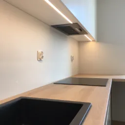 Nowoczesna kuchnia z drewnianym blatem, czarnym zlewem i płytą indukcyjną, oświetlenie LED podszafkowe, minimalistyczny okap, gniazdka elektryczne na ścianie.