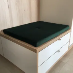 Siedzisko z ciemnozielonego weluru z guzikiem, umieszczone na szafce z białymi frontami szuflad i blatem z jasnego drewna, wkomponowane we wnękę z panelami ściennymi.