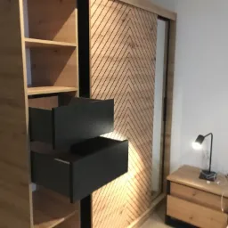 Szafa z otwartymi szufladami w kolorze grafitowym, drewniane półki i przesuwne drzwi z wzorem w jodełkę, obok szafka nocna z lampką.