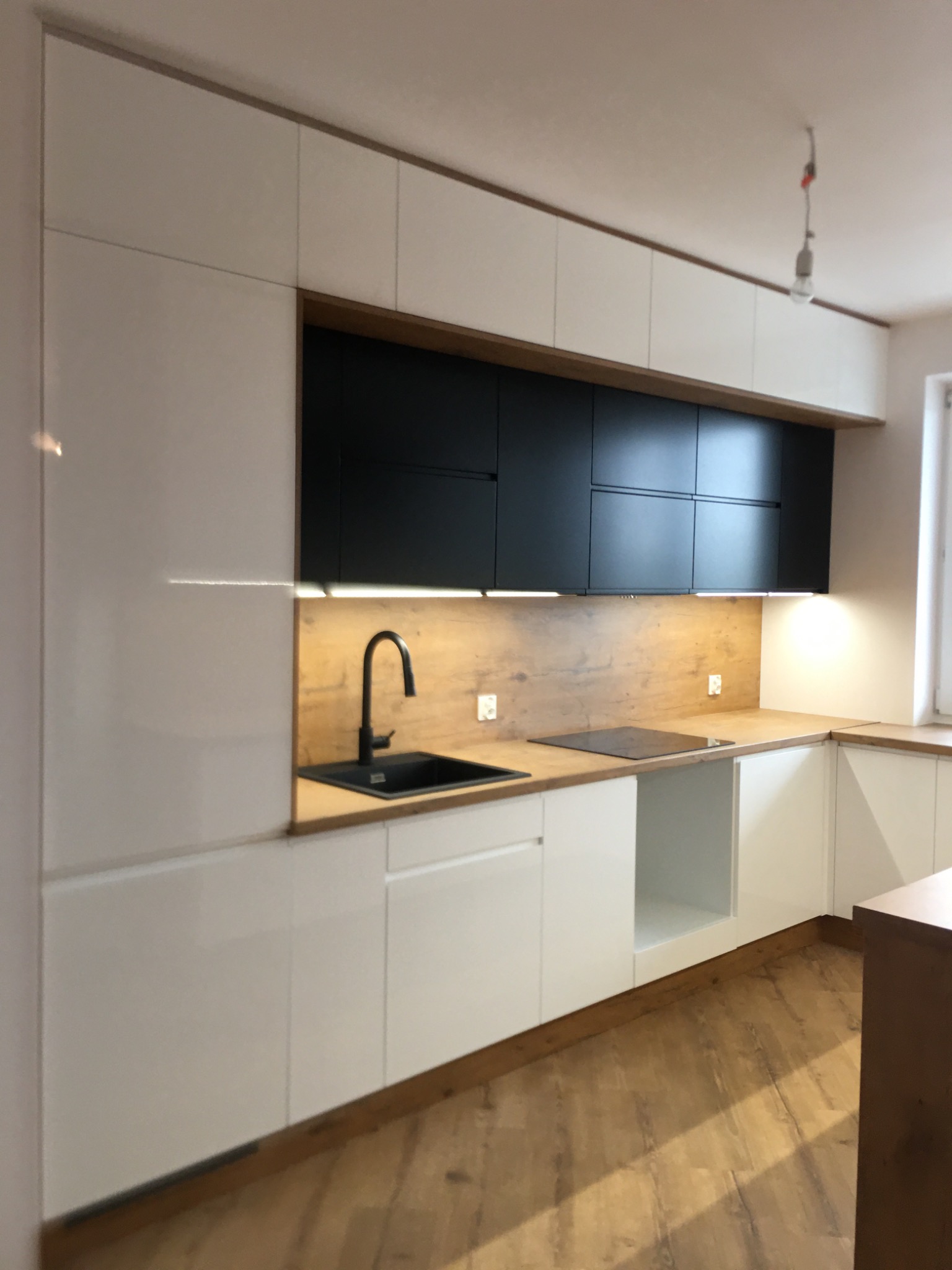Nowoczesna kuchnia na wymiar z białymi frontami dolnymi i czarnymi górnymi szafkami, drewnianym blatem i oświetleniem LED podszafkowym, widok na zlew i płytę indukcyjną.