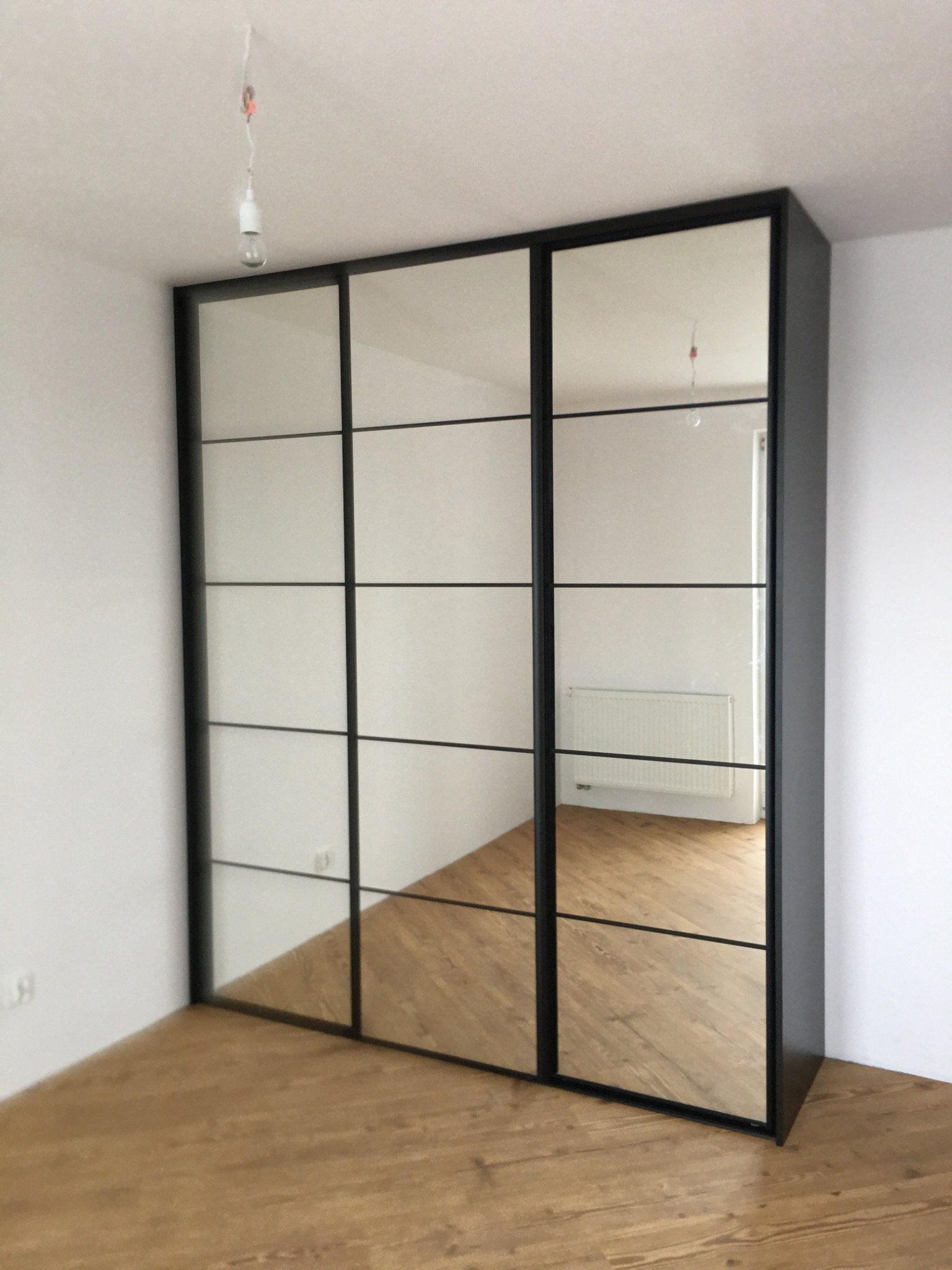 Trzydrzwiowa szafa wnękowa z lustrami i czarnymi szprosami, widoczna na tle białych ścian i drewnianej podłogi; w lustrach odbija się grzejnik i fragment drugiego pokoju.