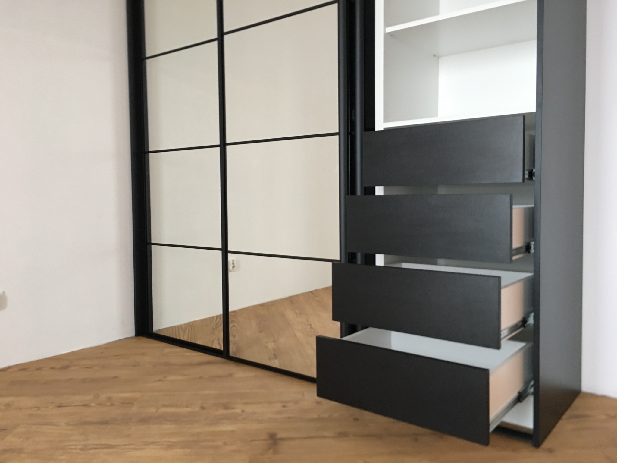 Czarna szafa z przesuwnymi drzwiami z podziałem na prostokątne pola i otwartymi szufladami, widoczna na tle drewnianej podłogi.