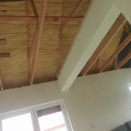 Poddasze w trakcie prac: widoczne drewniane krokwie, izolacja z wełny mineralnej, fragment ściany po nałożeniu pierwszej warstwy gładzi, okno dachowe.