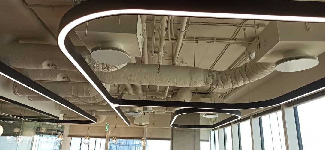 Biały sufit biurowy z widocznymi elementami instalacji, takimi jak rury, przewody i kratki wentylacyjne, częściowo otulone izolacją, oświetlony nowoczesnymi lampami LED o geometrycznych kształtach.